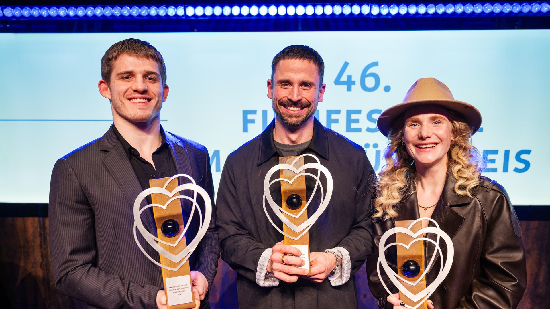 Regisseur Lauro Cress (M) steht auf der Bühne. Er hält eine Trophäe in der Hand. Neben ihm stehen die Schauspielerin Ladina von Frisching und der Schauspieler Giulio Brizzi. Auch sie halten jeweils eine Trophäe in den Händen.