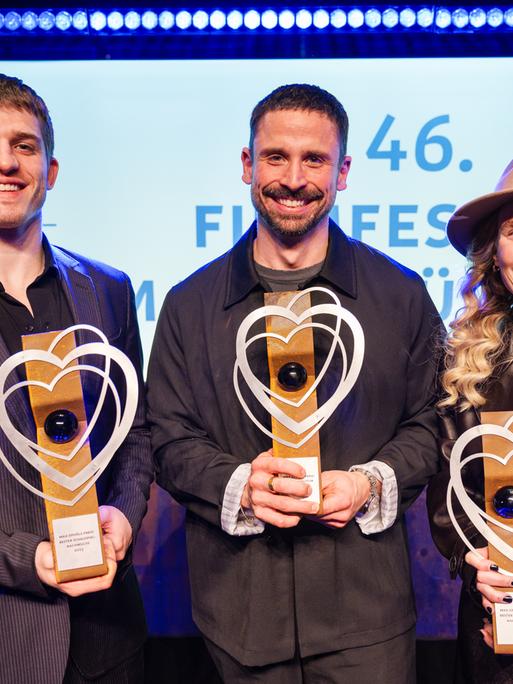 Regisseur Lauro Cress (M) steht auf der Bühne. Er hält eine Trophäe in der Hand. Neben ihm stehen die Schauspielerin Ladina von Frisching und der Schauspieler Giulio Brizzi. Auch sie halten jeweils eine Trophäe in den Händen.