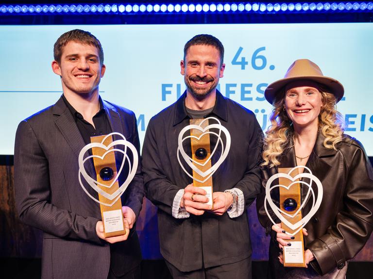 Regisseur Lauro Cress (M) steht auf der Bühne. Er hält eine Trophäe in der Hand. Neben ihm stehen die Schauspielerin Ladina von Frisching und der Schauspieler Giulio Brizzi. Auch sie halten jeweils eine Trophäe in den Händen.