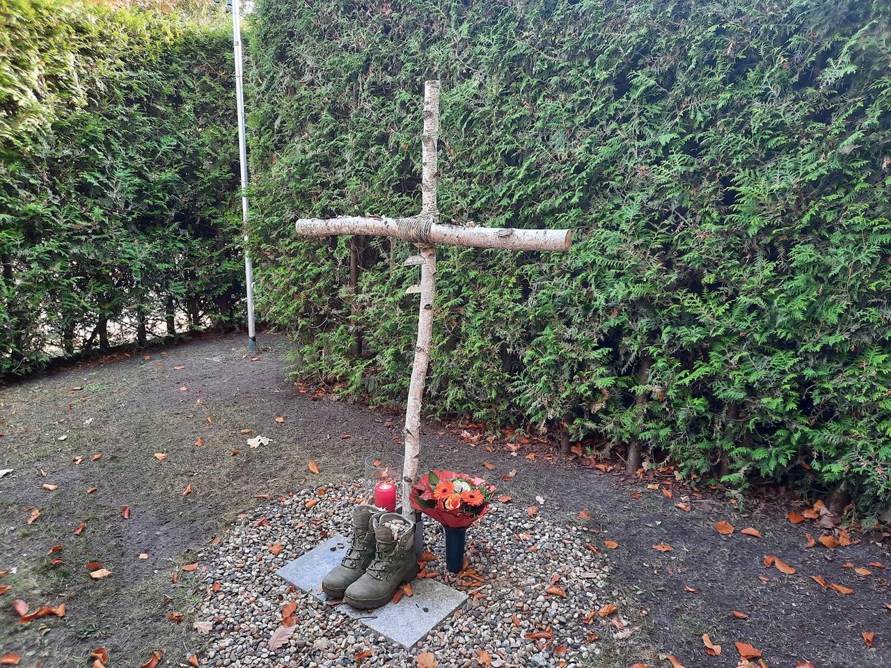 Vor einem Holzkreuz steht ein Paar robuste Stiefel, befüllt mit Kieselsteinen. Daneben steht eine Kerze und ein Vase mit Blumen.