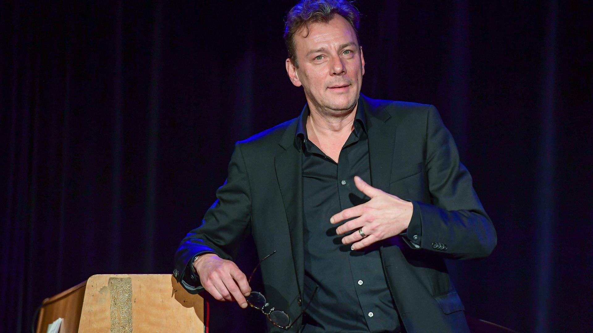 Alexander Suckel, Leiter des Literaturhauses Halle, lehnt sich an ein Klavier und gestikuliert. 
