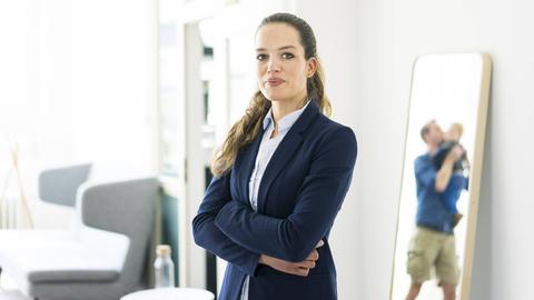 Eine Frau mit Business-Outfit steht neben einem Spiegel, in dem ein Mann mit Kleinkind zu sehen ist.