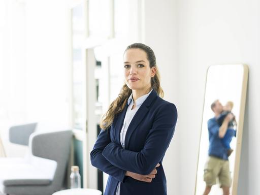 Eine Frau mit Business-Outfit steht neben einem Spiegel, in dem ein Mann mit Kleinkind zu sehen ist.