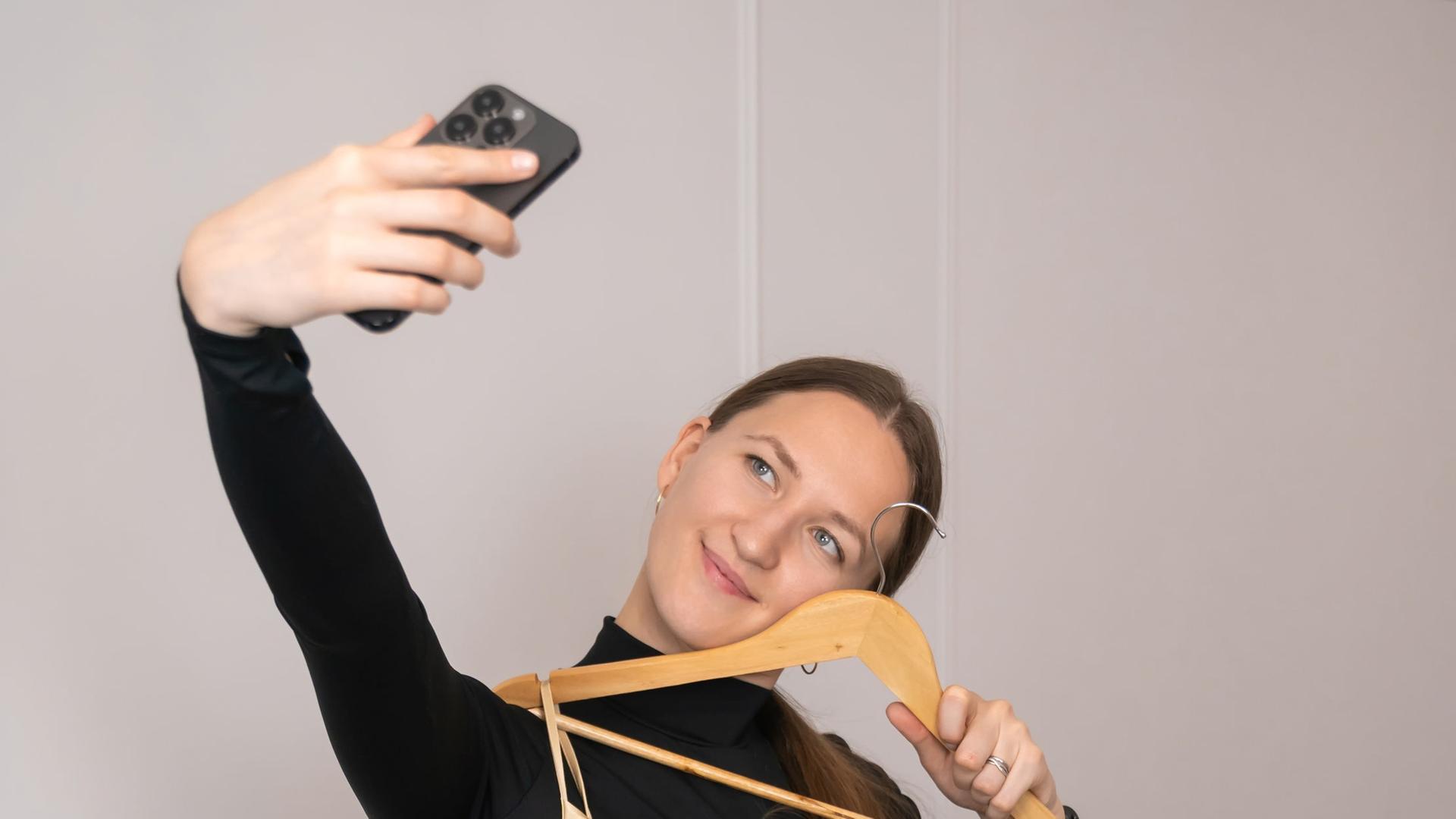 Eine junge Frau hält ein langes Kleid auf einem Kleiderbügel vor ihren Körper, während sie mit ihrem Smartphone ein Selfie macht.