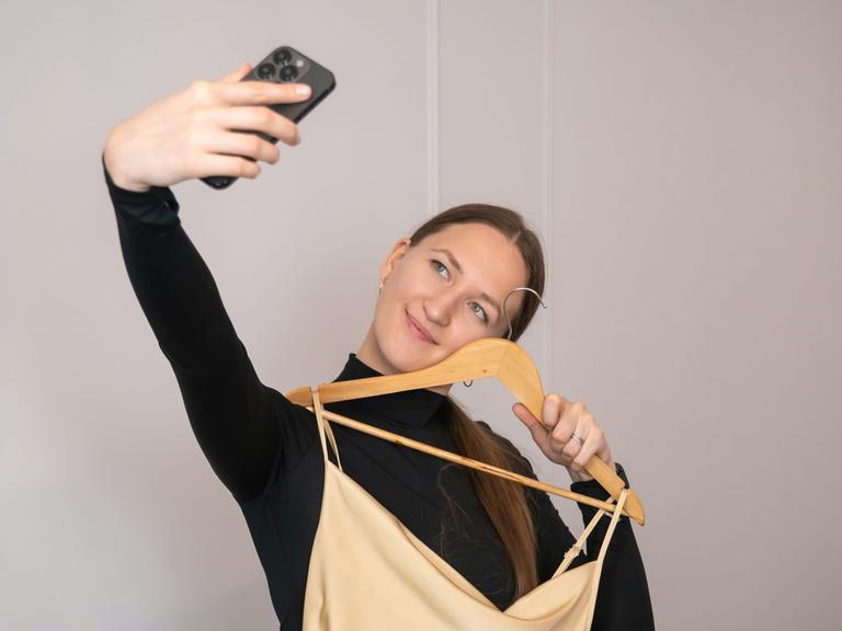 Eine junge Frau hält ein langes Kleid auf einem Kleiderbügel vor ihren Körper, während sie mit ihrem Smartphone ein Selfie macht.