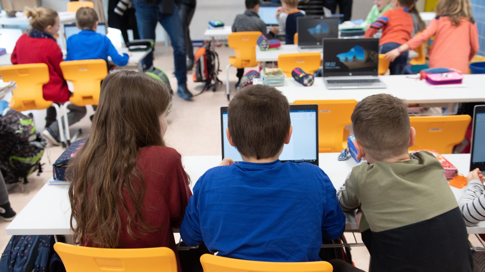 Drei Grundschüler sitzen in einem vollen Klassenzimmer vor einem Laptop.