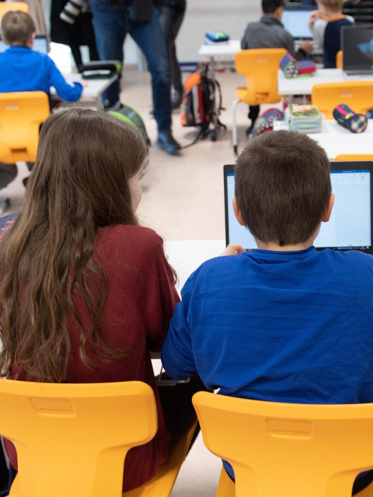 Drei Grundschüler sitzen in einem vollen Klassenzimmer vor einem Laptop.