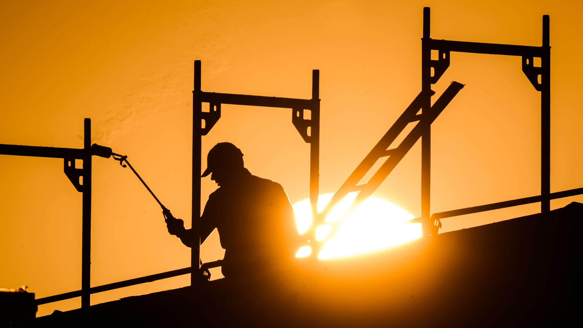 Ein Bauarbeiter steht mit einem Gasbrenner beim Bau eines Mehrfamilienhauses in einem Neubaugebiet als am Horizont die Sonne aufgeht.
