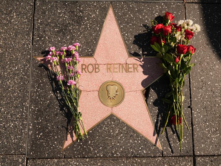 Nahaufnahme des Sterns auf dem Bürgersteig, an dem rechts und links zwei Blumenstäuße liegen. 