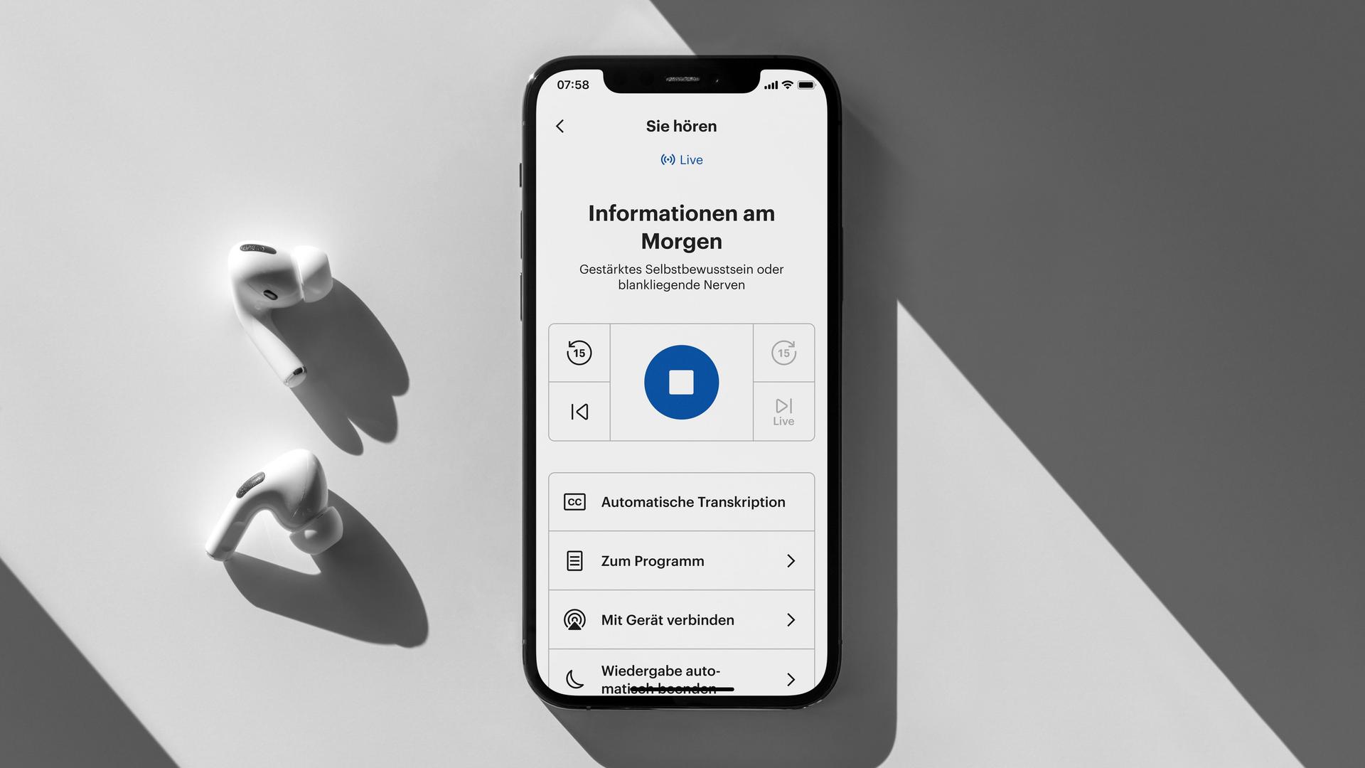 Ein Smartphone mit der geöffneten Deutschlandfunk App im einfachen Modus und zwei kabellose In-Ear Kopfhörer liegen auf einem hellen Schreibtisch.