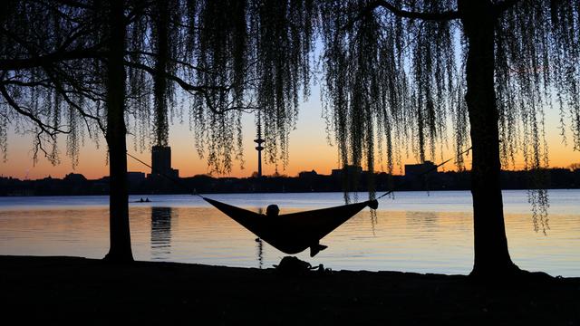 Ein Mensch liegt an einem See in einer Hängematte. Im Hintergrund sind Gebäude erkennbar. Die Sonne geht gerade unter.