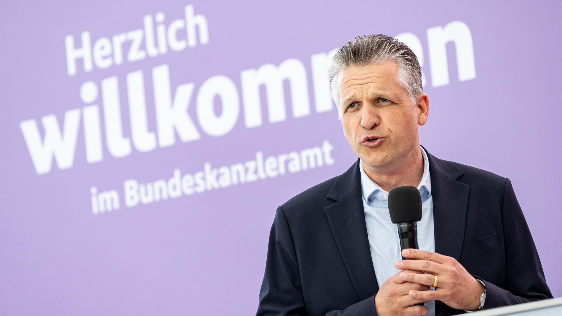 Frei hält ein Mikrofon in beiden Händen und scheint jemandem zuzuhören. Dahinter eine lilafarbene Wand. Darauf steht mit weißer Schrift: "Herzlich willkommen im Bundeskanzleramt". Frei hält ein Mikrofon in beiden Händen und scheint jemandem zuzuhören. Dahinter eine lilafarbene Wand. Darauf steht mit weißer Schrift: "Herzlich willkommen im Bundeskanzleramt".