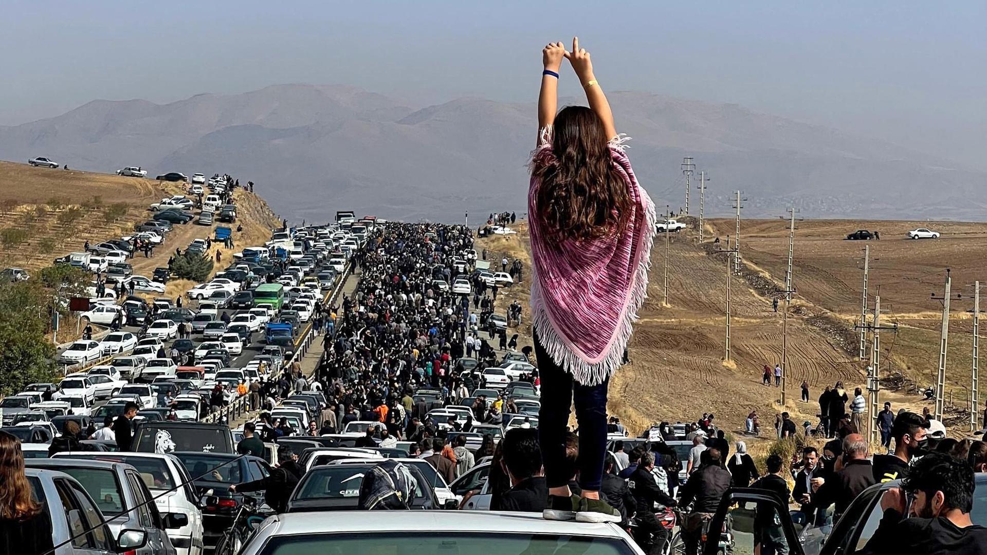 Das Foto zeigt eine Demonstration im Iran. Vorne steht eine Frau ohne Kopf-Tuch auf einem Auto.
