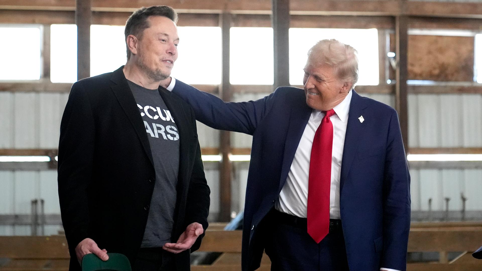 Donald Trump legt seine Hand aus die Schulter von Elon Musk und lächelt ihn an, während die beiden in einem Stall stehen.