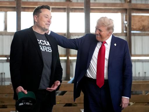 Donald Trump legt seine Hand aus die Schulter von Elon Musk und lächelt ihn an, während die beiden in einem Stall stehen.