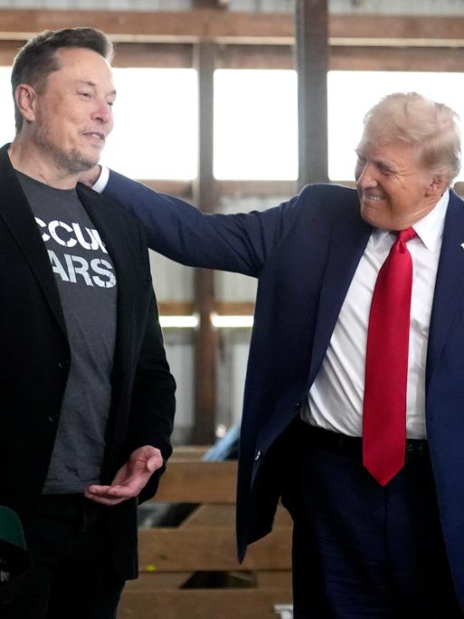 Donald Trump legt seine Hand aus die Schulter von Elon Musk und lächelt ihn an, während die beiden in einem Stall stehen.