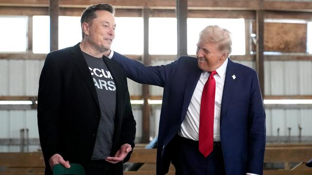 Elon Musk steht neben Donald Trump. Trump legt seine Hand auf die Schulter von Musk. Beide grinsen.