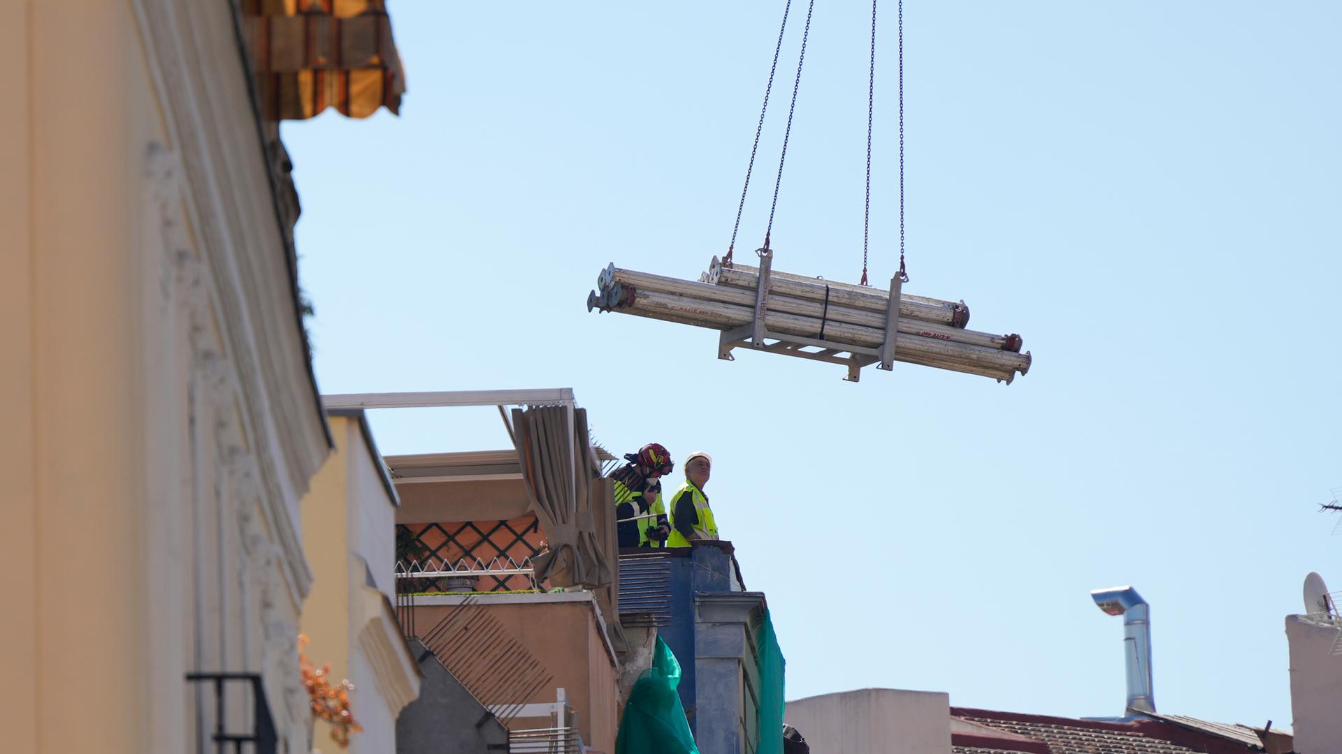 Blick aus größerer Entfernung: Zwei Männer in gelben Warnwesten und mit Helmen stehen am Dachrand. An der Kette eines Krans hängen mehrere Rohre. Darüber blauer Himmel.