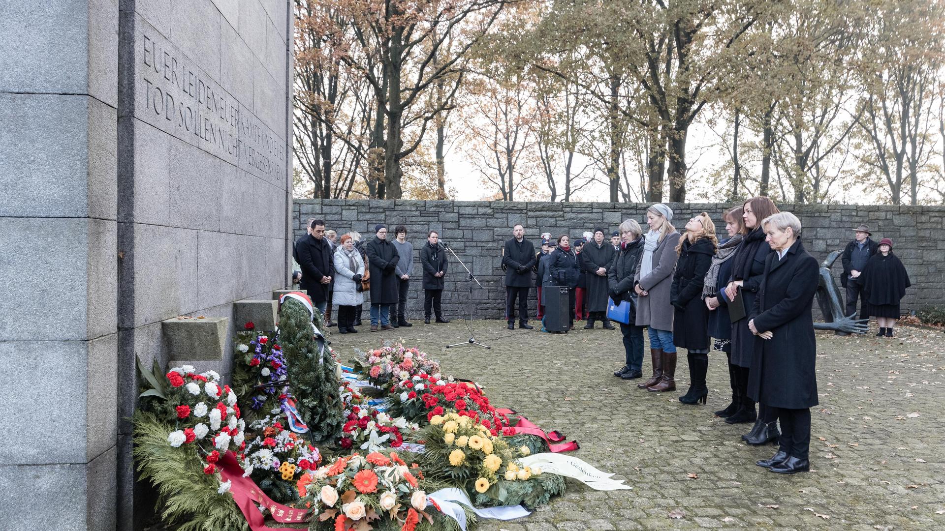 An einer Gedenkmauer lehnen mehrere Blumenkränze. Sechs Frauen stehen vor den Kränzen. Mehrere Menschen stehen um die Frauen herum