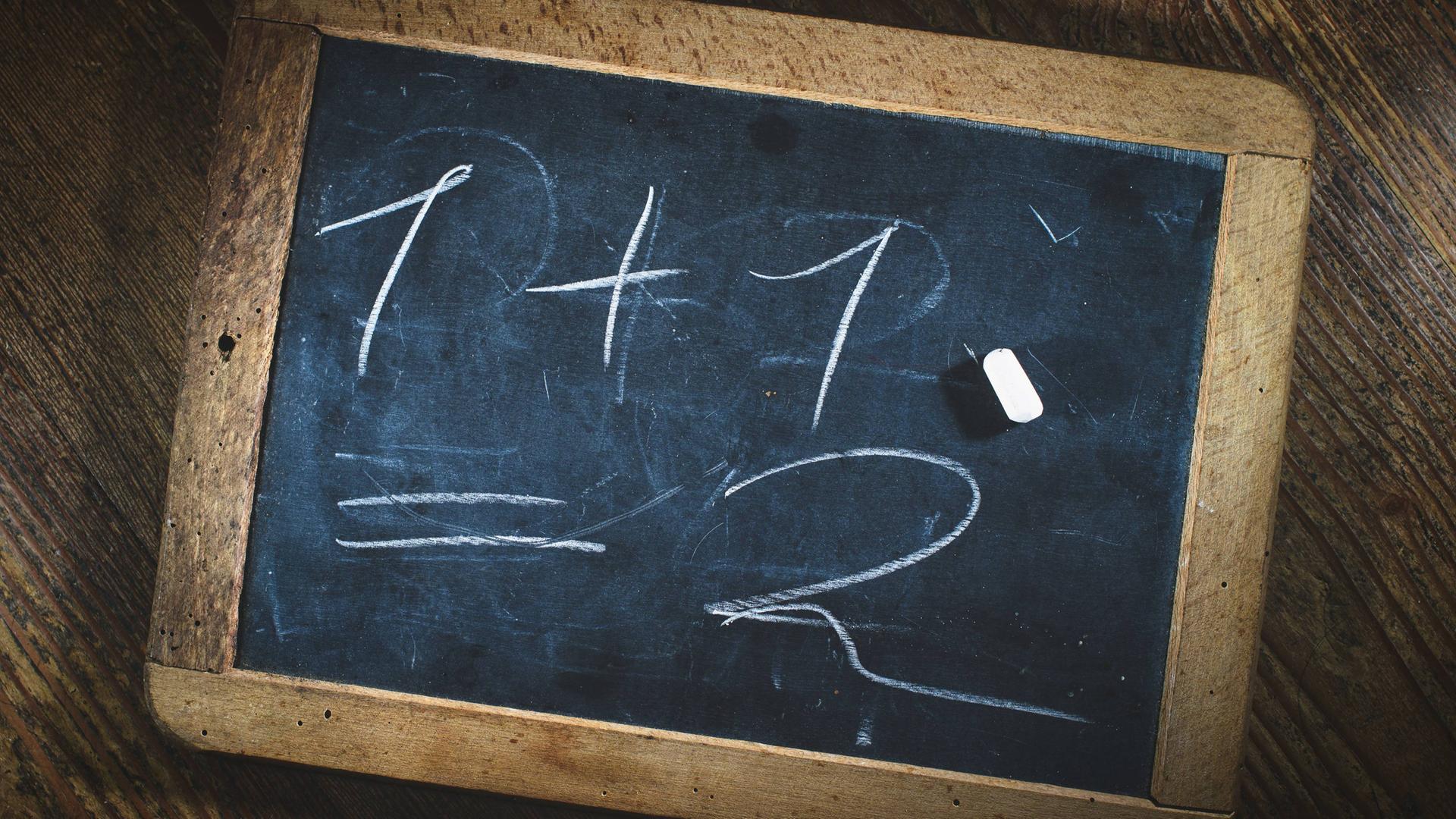 Eine kleine Kreidetafel, auf der steht: 1 +1 = 2. Ein Stück Kreide liegt darauf. Eine kleine Kreidetafel, auf der steht: 1 +1 = 2. Ein Stück Kreide liegt darauf.