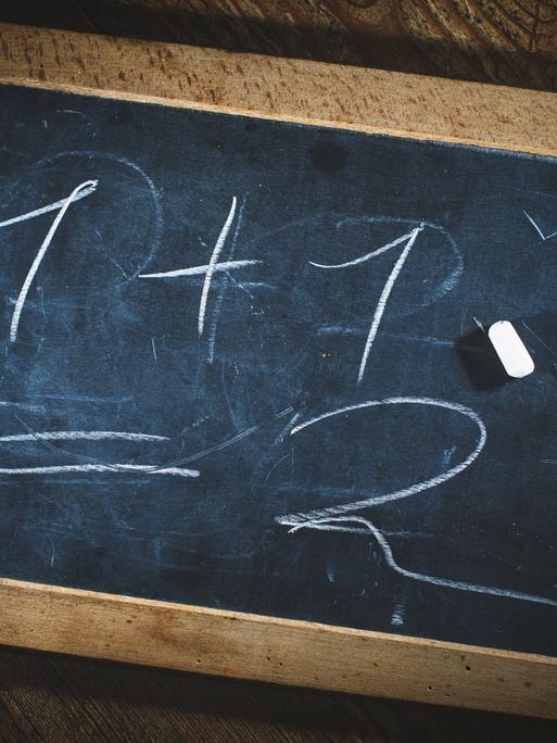 Eine kleine Kreidetafel, auf der steht: 1 +1 = 2. Ein Stück Kreide liegt darauf.