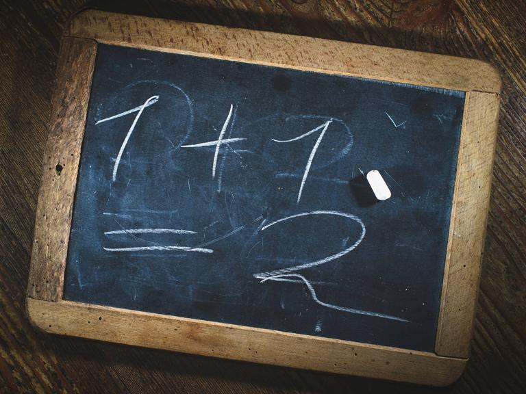Eine kleine Kreidetafel, auf der steht: 1 +1 = 2. Ein Stück Kreide liegt darauf.