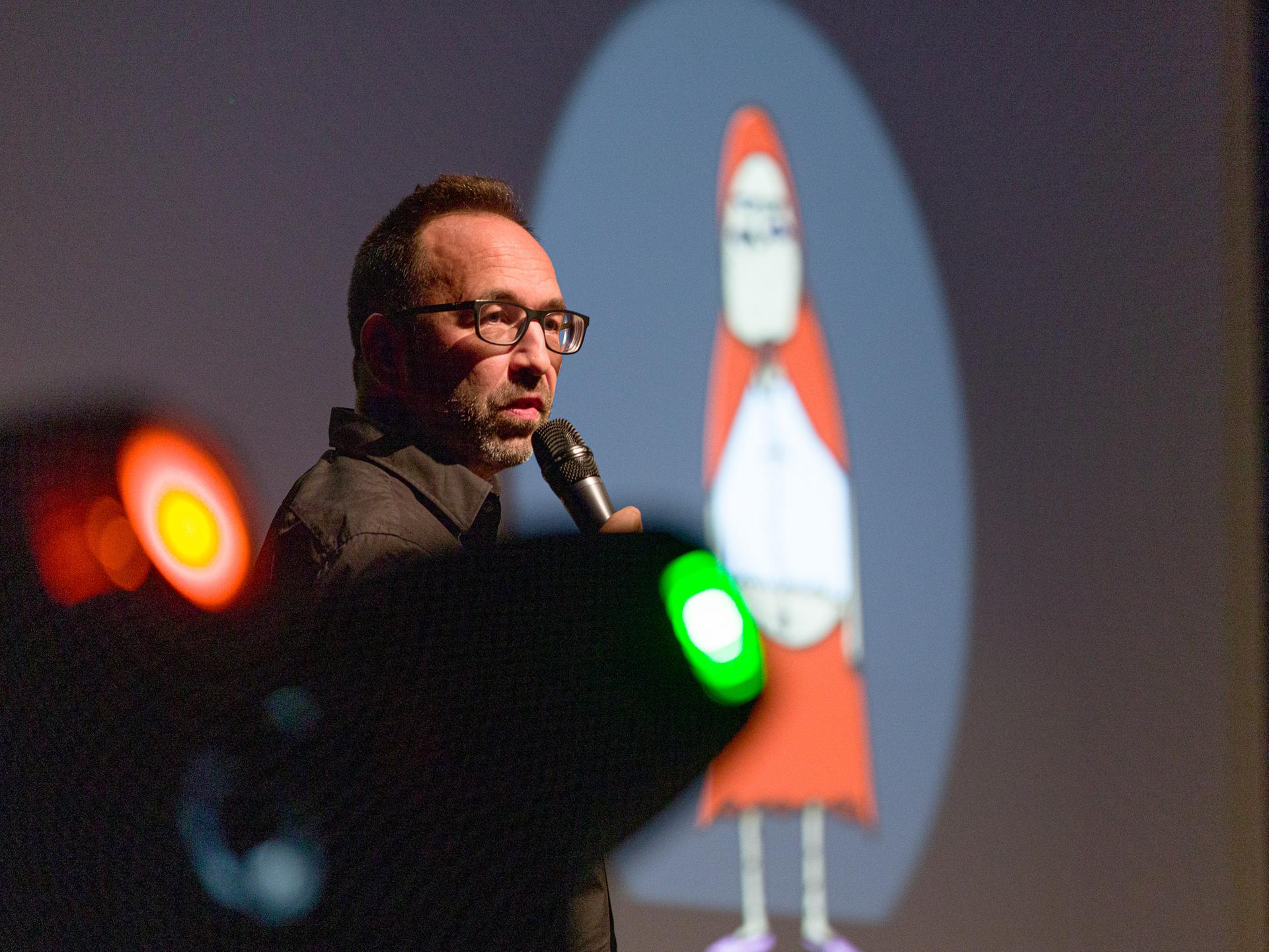 Piero Masztalerz steht beim ComedyArts-Festival 2022 in Moers mit einem Mikrofon auf der Bühne. Im Hintergrund ist ein Weihnachtsmann-Cartoon von ihm zu sehen.