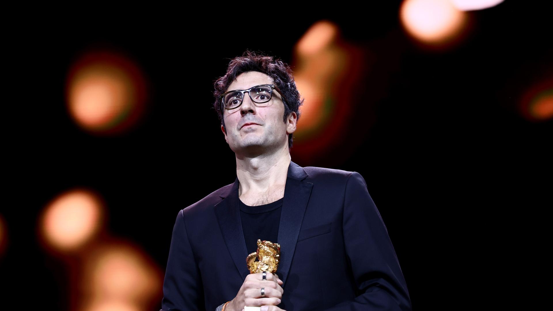Das Bild zeigt den Regisseur Ilker Çatak im dunklen Anzug auf einer Bühne. Er schaut ehrfürchtig ins Publikum und hält den Goldenen Bären in den Händen. Im Hintergrund sind verschwommene Lichter zu erkennen.