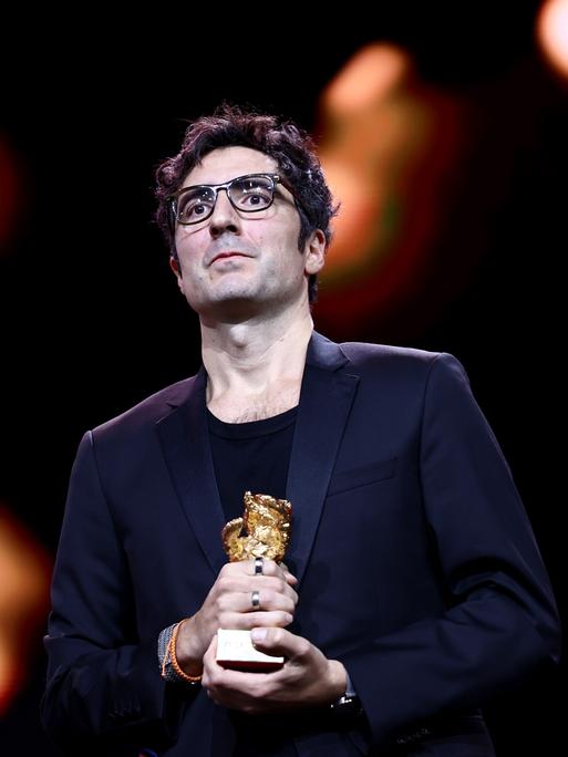 Das Bild zeigt den Regisseur Ilker Çatak im dunklen Anzug auf einer Bühne. Er schaut ins Publikum und hält den Goldenen Bären in den Händen. Im Hintergrund sind verschwommene Lichter.