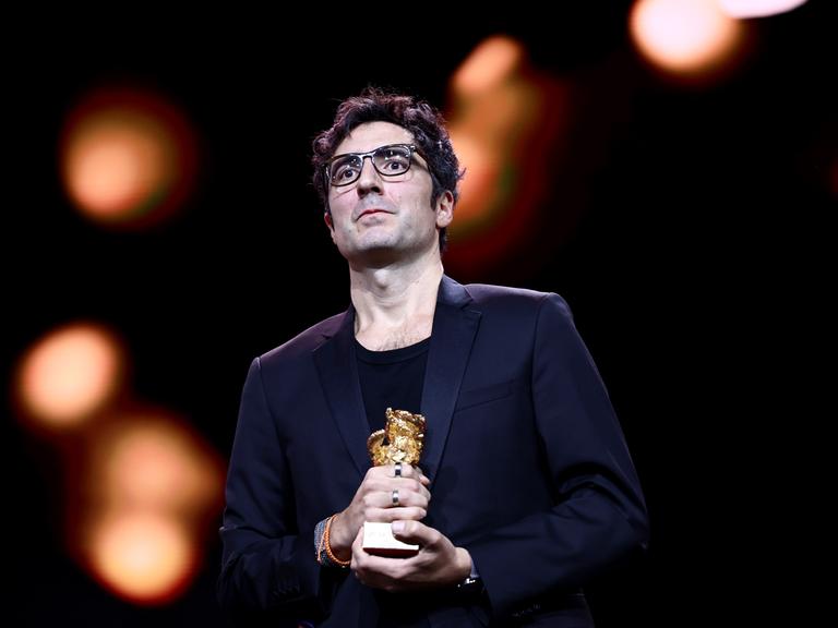 Das Bild zeigt den Regisseur Ilker Çatak im dunklen Anzug auf einer Bühne. Er schaut ins Publikum und hält den Goldenen Bären in den Händen. Im Hintergrund sind verschwommene Lichter.