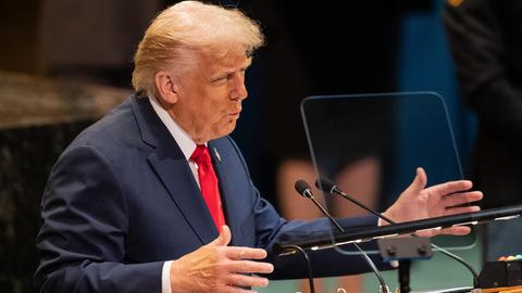 US-Präsident Trump steht in einem blauen Anzug mit einer roten Krawatte an einem Rednerpult. Er spricht und gestikuliert mit seinen Händen.