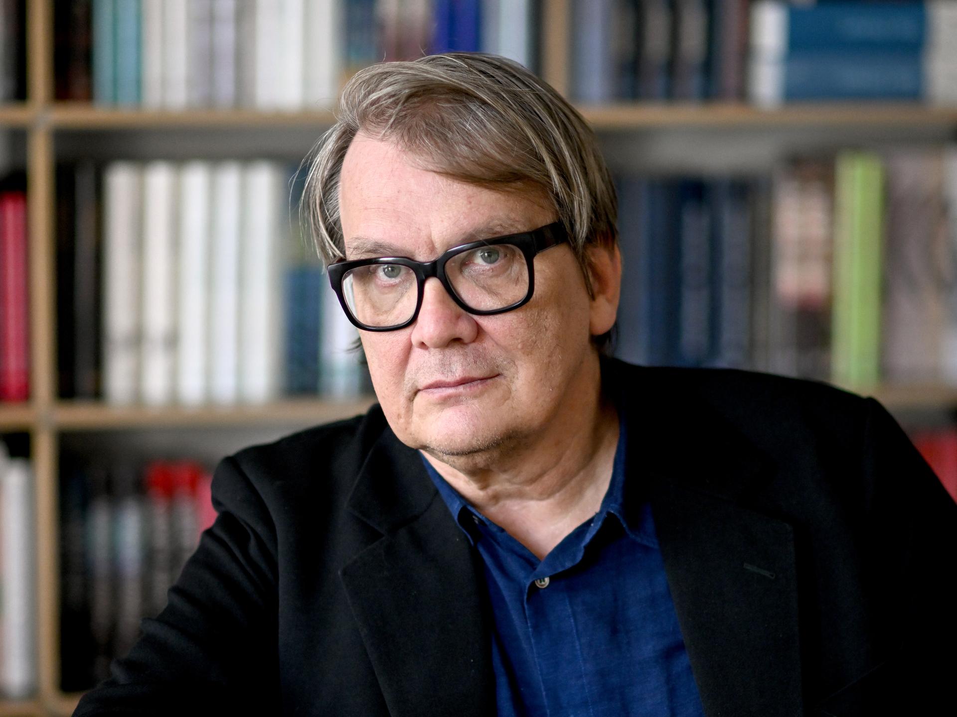 Der Schriftsteller Sven Regener im Portrait. Er trägt eine dicke, schwarze Brille ein blaues Hemd und eine dunkle Jacke. Hinter ihm ist ein Bücherregal. Der Schriftsteller Sven Regener im Portrait. Er trägt eine dicke, schwarze Brille ein blaues Hemd und eine dunkle Jacke. Hinter ihm ist ein Bücherregal.