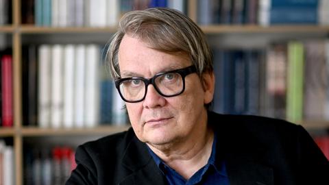 Der Schriftsteller Sven Regener im Portrait. Er trägt eine dicke, schwarze Brille ein blaues Hemd und eine dunkle Jacke. Hinter ihm ist ein Bücherregal. Der Schriftsteller Sven Regener im Portrait. Er trägt eine dicke, schwarze Brille ein blaues Hemd und eine dunkle Jacke. Hinter ihm ist ein Bücherregal.