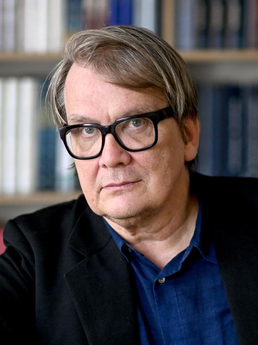 Der Schriftsteller Sven Regener im Portrait. Er trägt eine dicke, schwarze Brille ein blaues Hemd und eine dunkle Jacke. Hinter ihm ist ein Bücherregal. Der Schriftsteller Sven Regener im Portrait. Er trägt eine dicke, schwarze Brille ein blaues Hemd und eine dunkle Jacke. Hinter ihm ist ein Bücherregal.