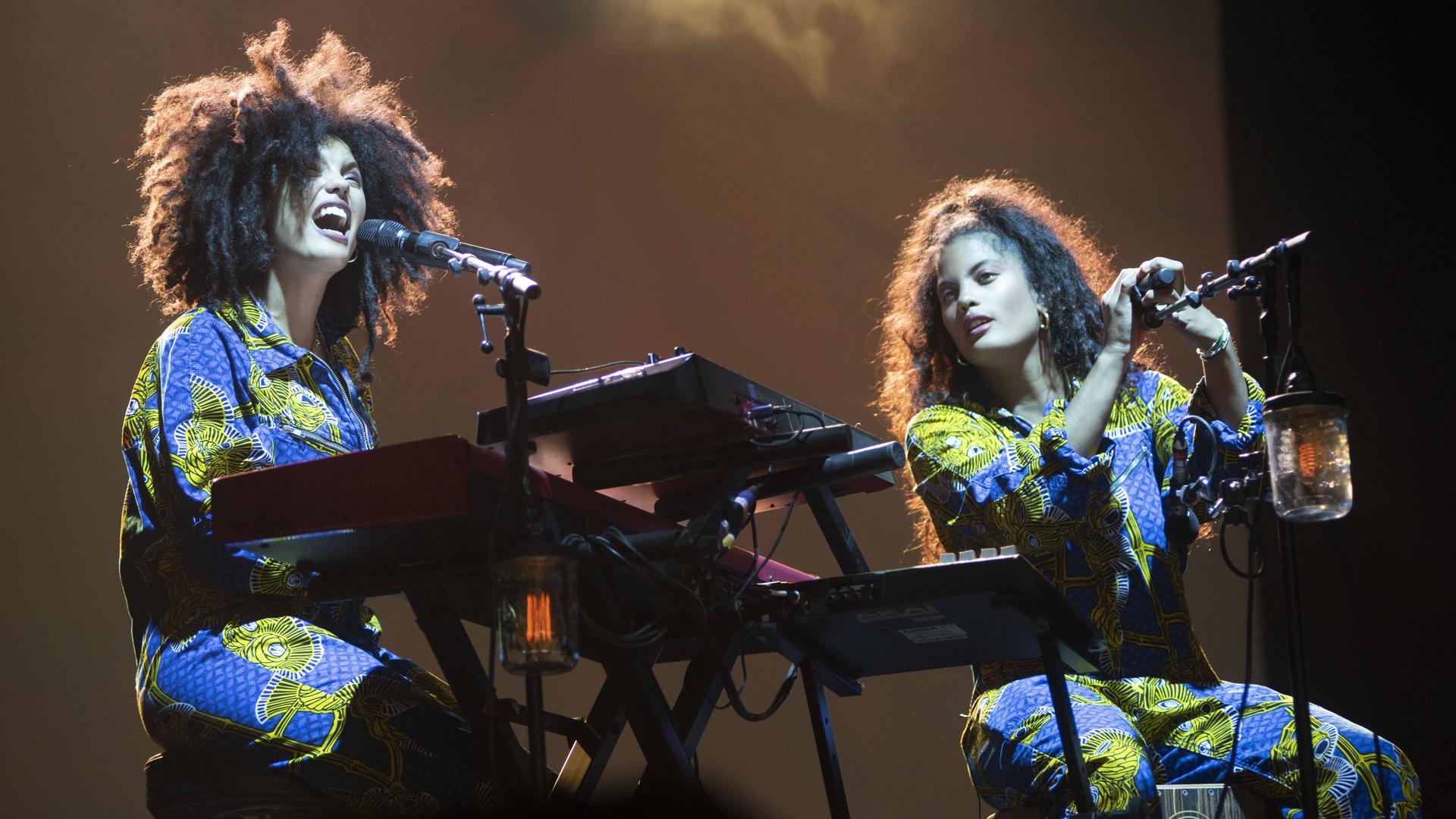 Die Zwillingsschwestern Lisa-Kainde Diaz und Naomi Diaz des Musikduos Ibeyi sitzen auf einer Bühne und singen. Das Licht ist schummerig, sie tragem Kleidung mit Afro-Mustern.