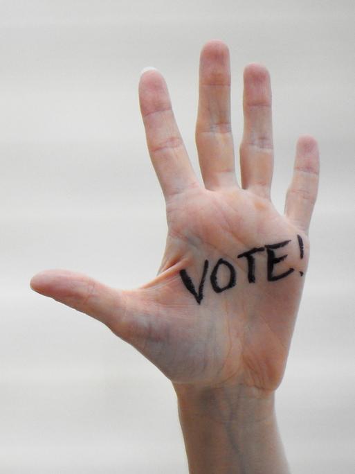 Eine ausgestrecke Hand wird in Großaufnahme gezeigt. Auf der Handinnenfläche ist in schwarzer Schrift "Vote" draufgeschrieben. Eine ausgestrecke Hand wird in Großaufnahme gezeigt. Auf der Handinnenfläche ist in schwarzer Schrift "Vote" draufgeschrieben.