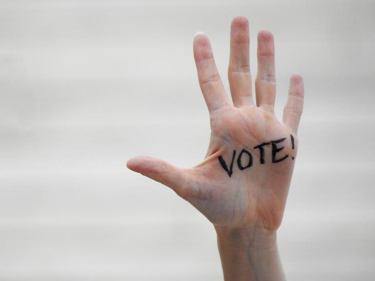Eine ausgestrecke Hand wird in Großaufnahme gezeigt. Auf der Handinnenfläche ist in schwarzer Schrift "Vote" draufgeschrieben. Eine ausgestrecke Hand wird in Großaufnahme gezeigt. Auf der Handinnenfläche ist in schwarzer Schrift "Vote" draufgeschrieben.