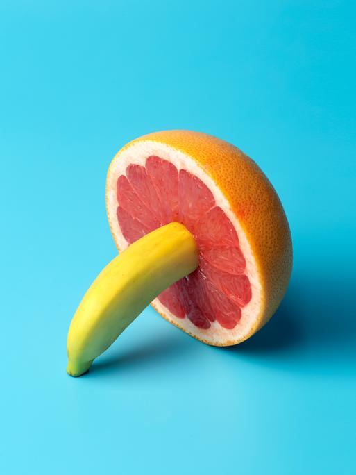 Minimalistische Darstellung einer Banane die in einer Grapefruit steckt auf blauem Grund.