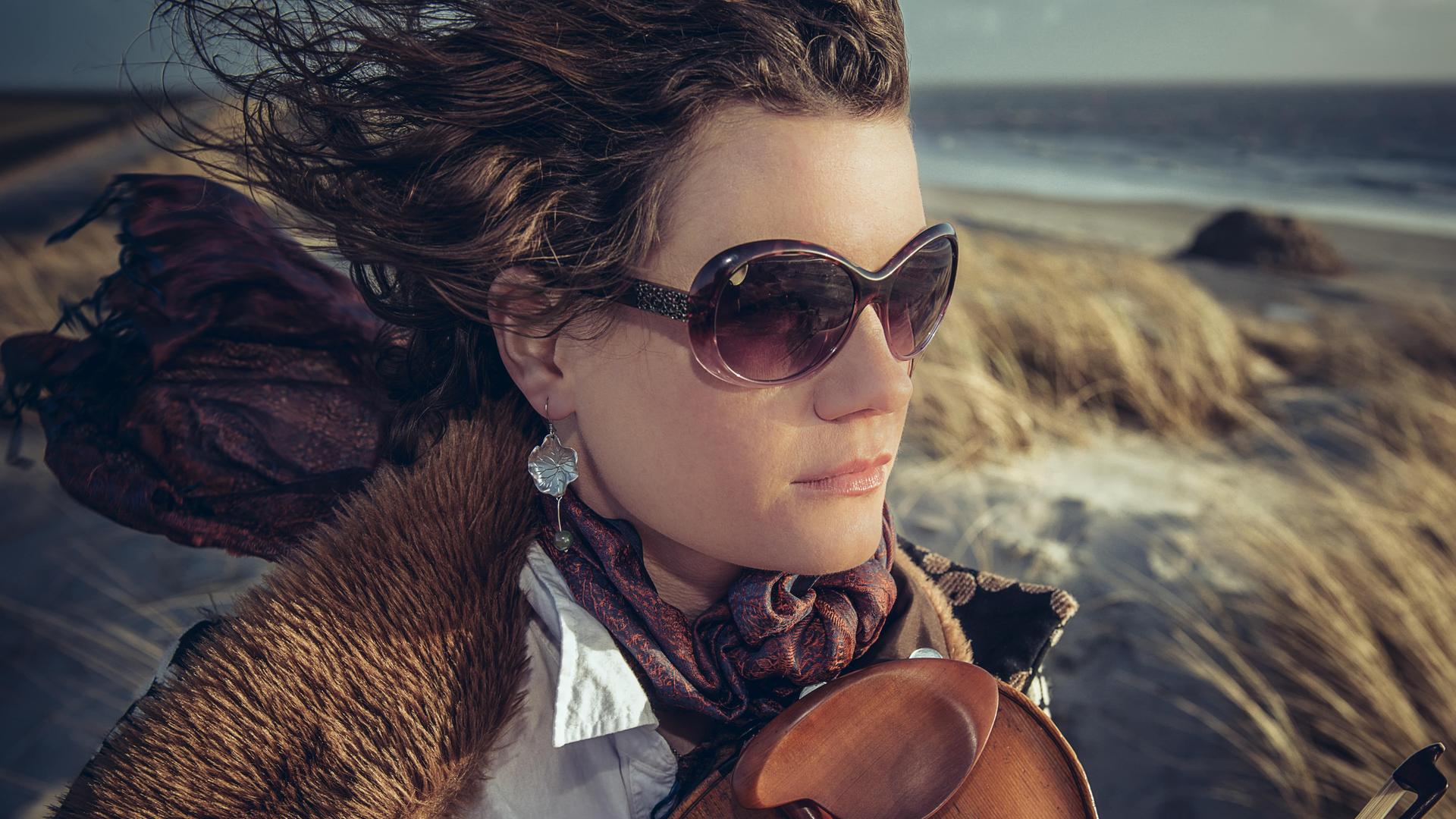 Eine Frau trägt Sonnenbrille und eine Jacke mit Pelzkragen über einer weißen Bluse. Der Wind weht ihre Haare und den buten Schal nach hinten. Auf ihrer Brust liegt locker eine Geige auf. Im Hintergrund sind Dünen und ein Meeresstrand zu sehen.