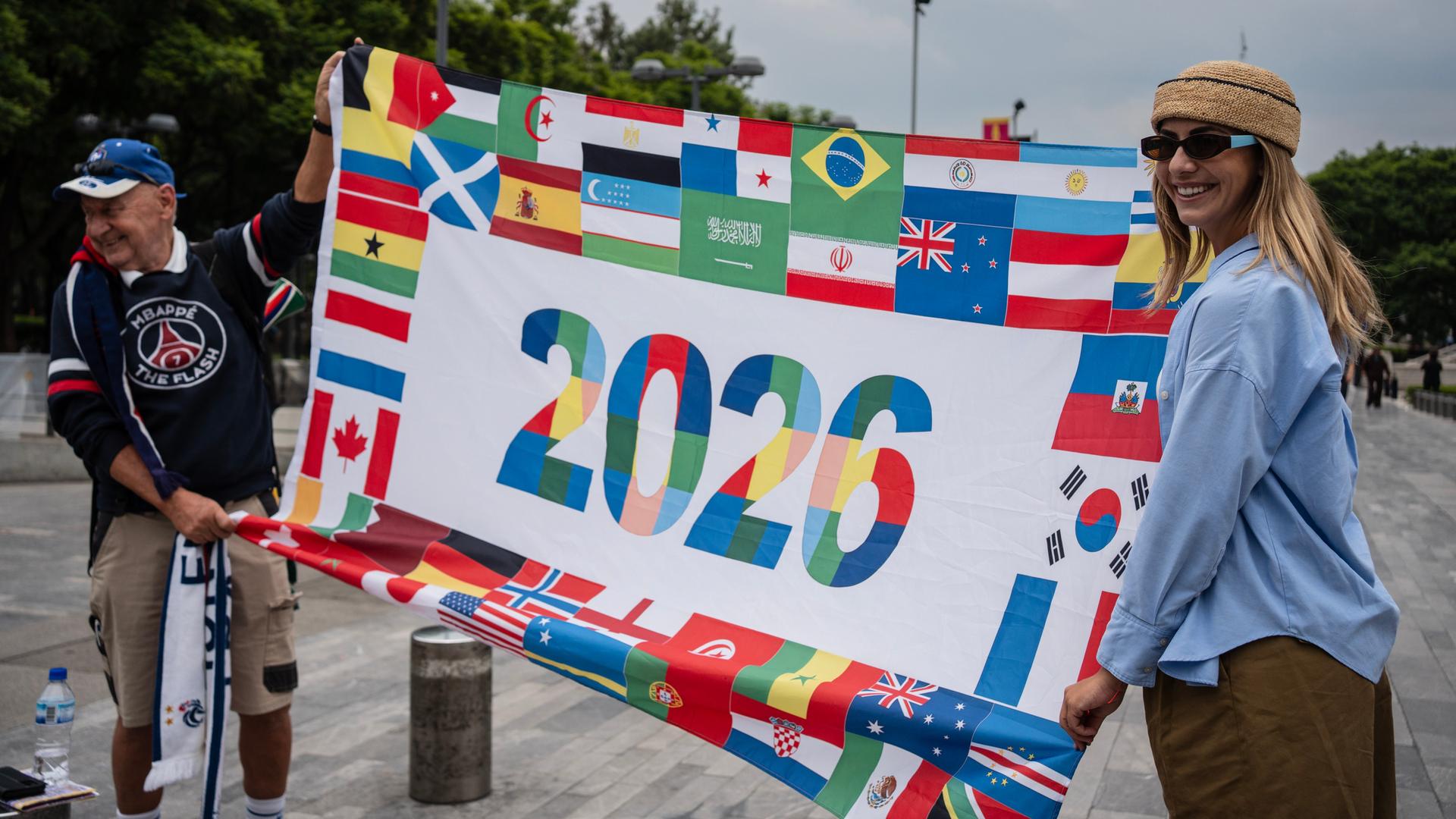 Französische Fußballfans posieren mit einem Transparent, auf dem um die Zahl 2026 die Flaggen aller WM-Teilnehmerländer angeordnet sind. Französische Fußballfans posieren mit einem Transparent, auf dem um die Zahl 2026 die Flaggen aller WM-Teilnehmerländer angeordnet sind.