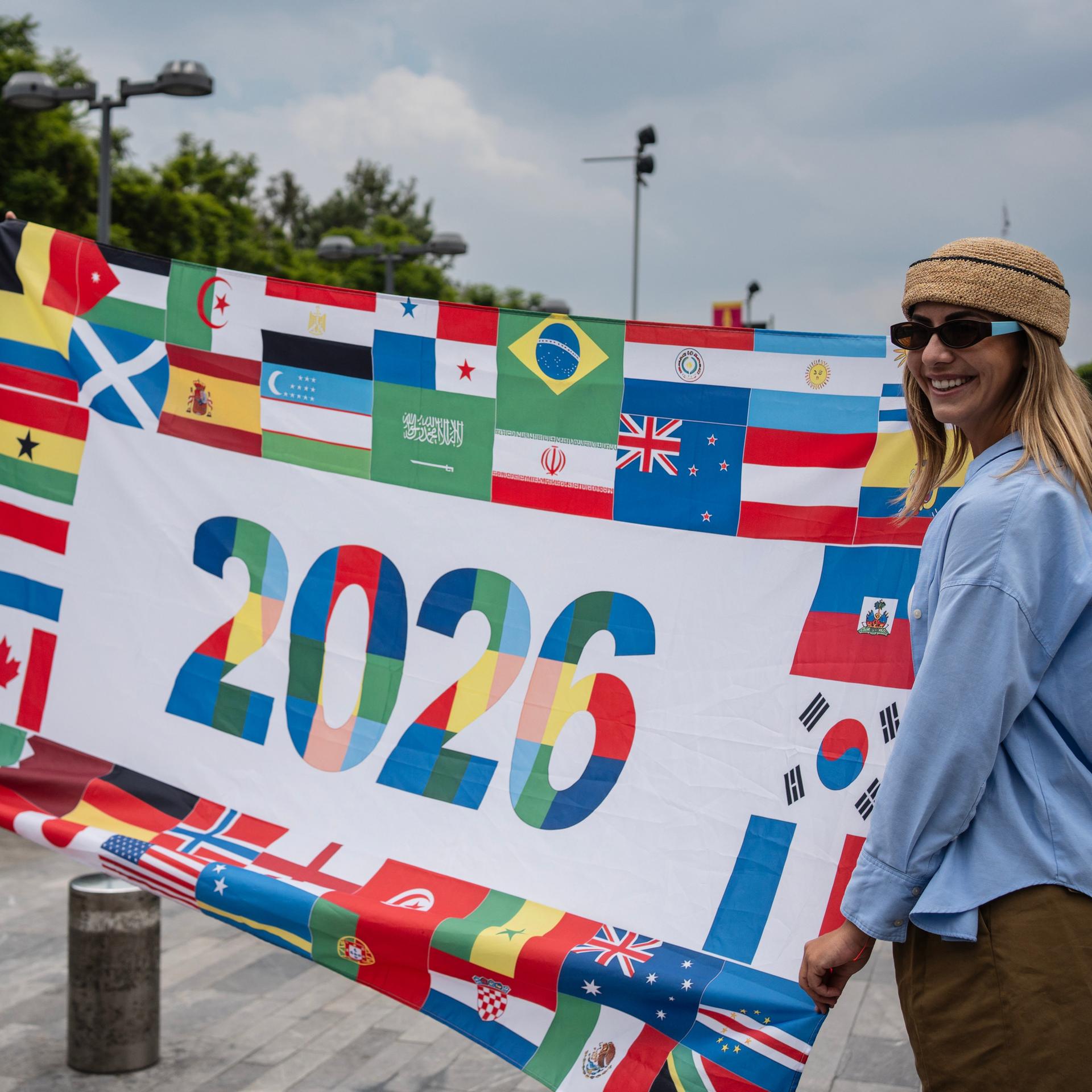 Französische Fußballfans posieren mit einem Transparent, auf dem um die Zahl 2026 die Flaggen aller WM-Teilnehmerländer angeordnet sind.