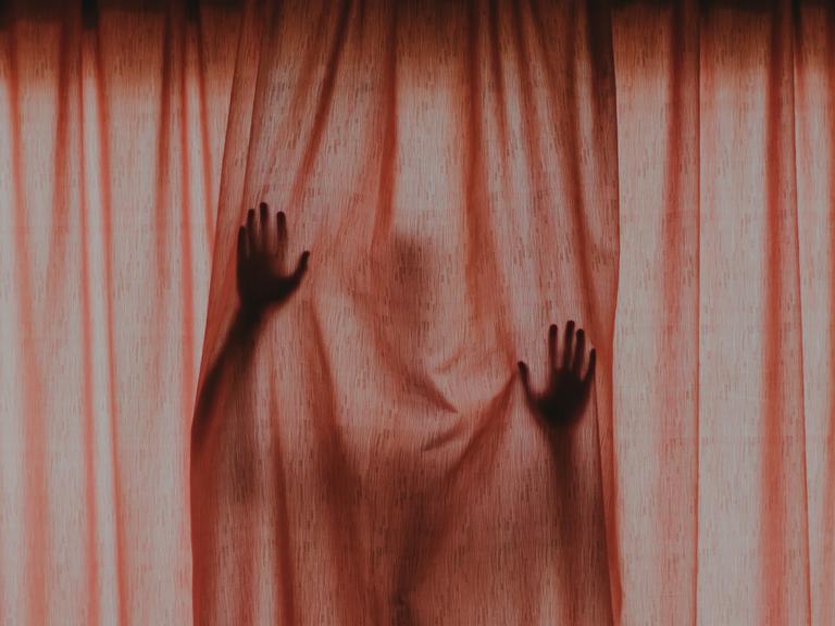 Eine Gestalt mit erhobenen Händen hinter einem verschlossenen roten Vorhang