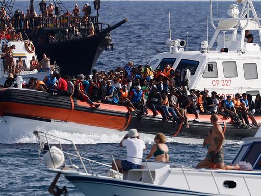Lampedusa im September 2023: Die italienische Küstenwache bringt Geflüchtete in den Hafen, während Touristin zuschauen.