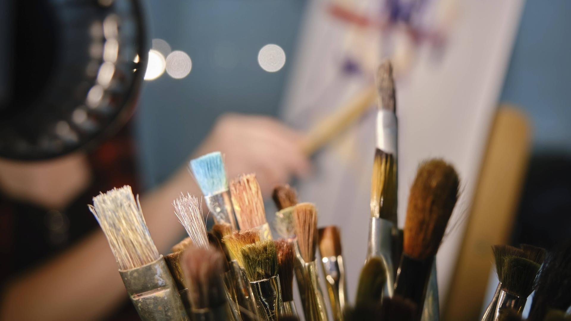 Behältnis mit Pinseln im Vordergrund. Im Hintergrund verschwommene Kunstschaffende in der Nähe Staffelei malt ein Bild. 