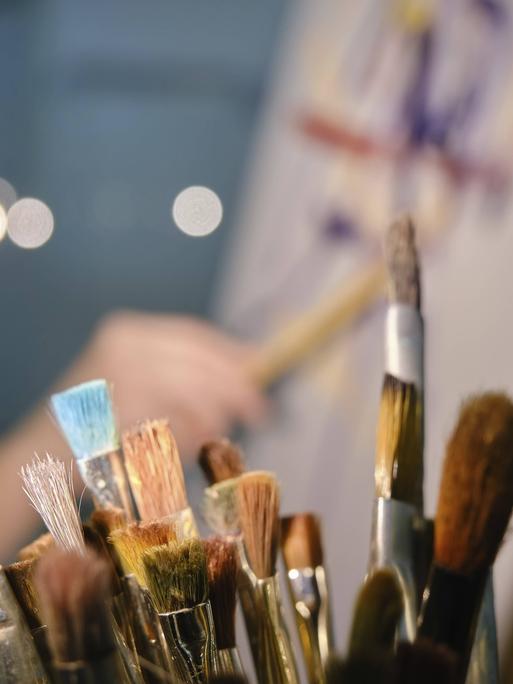Behältnis mit Pinseln im Vordergrund. Im Hintergrund verschwommene Kunstschaffende in der Nähe Staffelei malt ein Bild. 