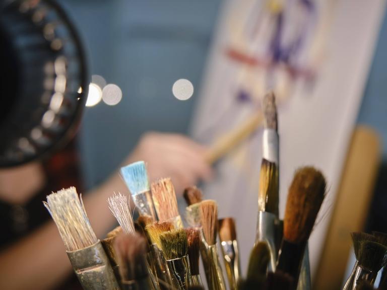 Behältnis mit Pinseln im Vordergrund. Im Hintergrund verschwommene Kunstschaffende in der Nähe Staffelei malt ein Bild. 