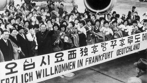 "Herzlich Willkommen in Frankfurt Deutschland" steht auf einem Spruchband, mit dem eine große Gruppe Krankenschwestern aus Südkorea im Jahr 1966 auf dem Rhein-Main-Flughafen begrüßt wird.