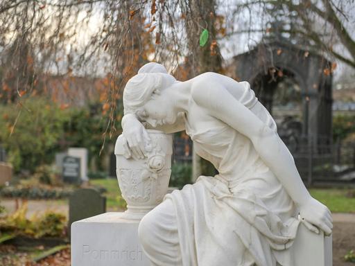 Die Skulptur auf dem Französischen Friedhof in Berlin zeigt eine sitzende Frauenfigur, die sich in Trauer versunken, mit geschlossenen Augen an eine neben ihr stehende Urne schmiegt und sich mit einer Hand an dem Stein festhält, auf dem sie sitzt. 