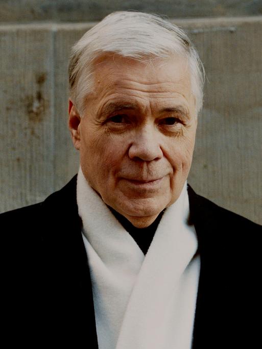 Dietrich Fischer-Dieskau steht in dunklem Mantel und mit weißem Schal vor einer Wand. Er hat weiße Haare und lächelt verschmitzt in die Kamera.