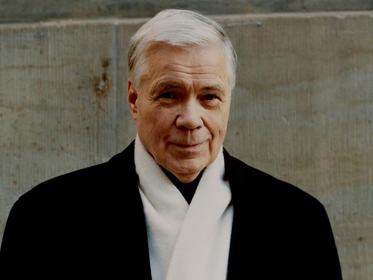 Dietrich Fischer-Dieskau steht in dunklem Mantel und mit weißem Schal vor einer Wand. Er hat weiße Haare und lächelt verschmitzt in die Kamera.